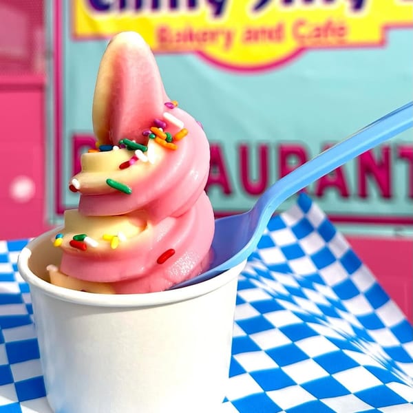 Swirled pink and yellow soft-serve ice cream with sprinkles in a white cup, on a blue checkered napkin.