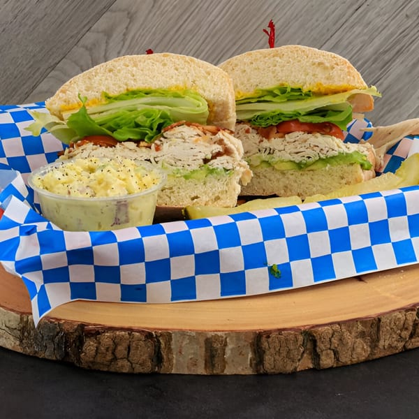 Chilly Jilly'z chicken sandwich with lettuce, tomato, and avocado, served with potato salad and pickles.
