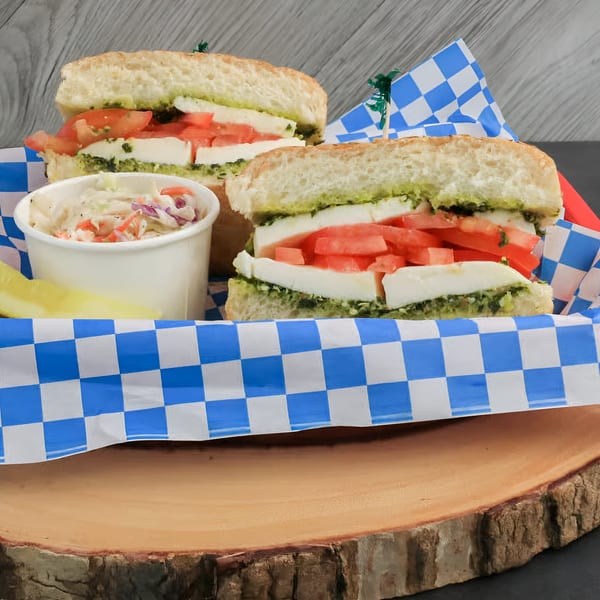 Two pesto, mozzarella, and tomato sandwiches with coleslaw and a pickle spear in a basket.
